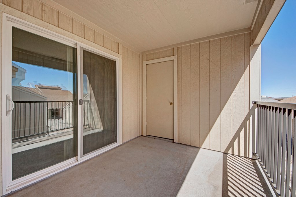 Small beige apartment balcony here at Fairgreen Apartment Homes with sliding glass door, wooden panel walls, and a simple metal railing casting shadows. Bright, sunny day outside.