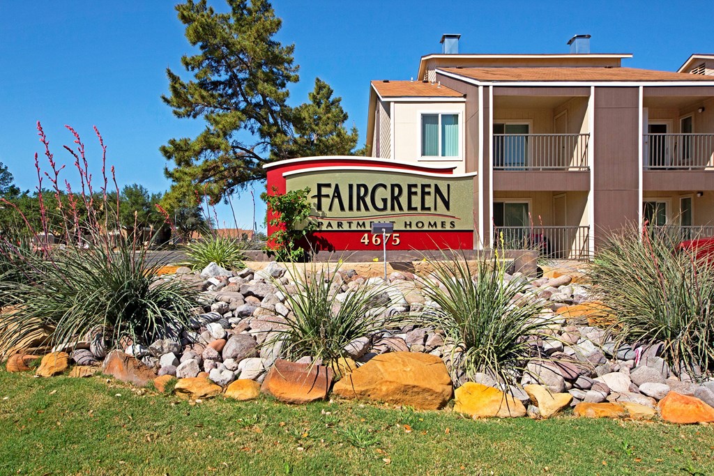Sign for Fairgreen Apartment Homes with address 4675, surrounded by greenery and rocks. Brown building in background under clear blue sky.