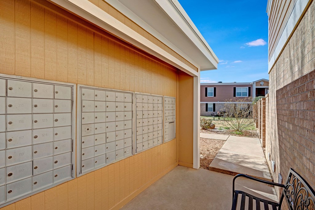 Row of mailboxes here at Golden Crest Apartment Homes in a sheltered area with a wooden bench nearby. Sunny day, blue sky, with residential buildings and pathway in the background.