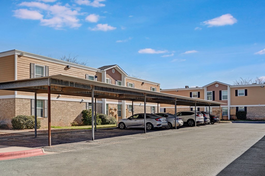 Golden Crest Apartment Homes with beige siding and brick walls under a clear blue sky. Covered parking area with several cars, casting shadows on the asphalt.