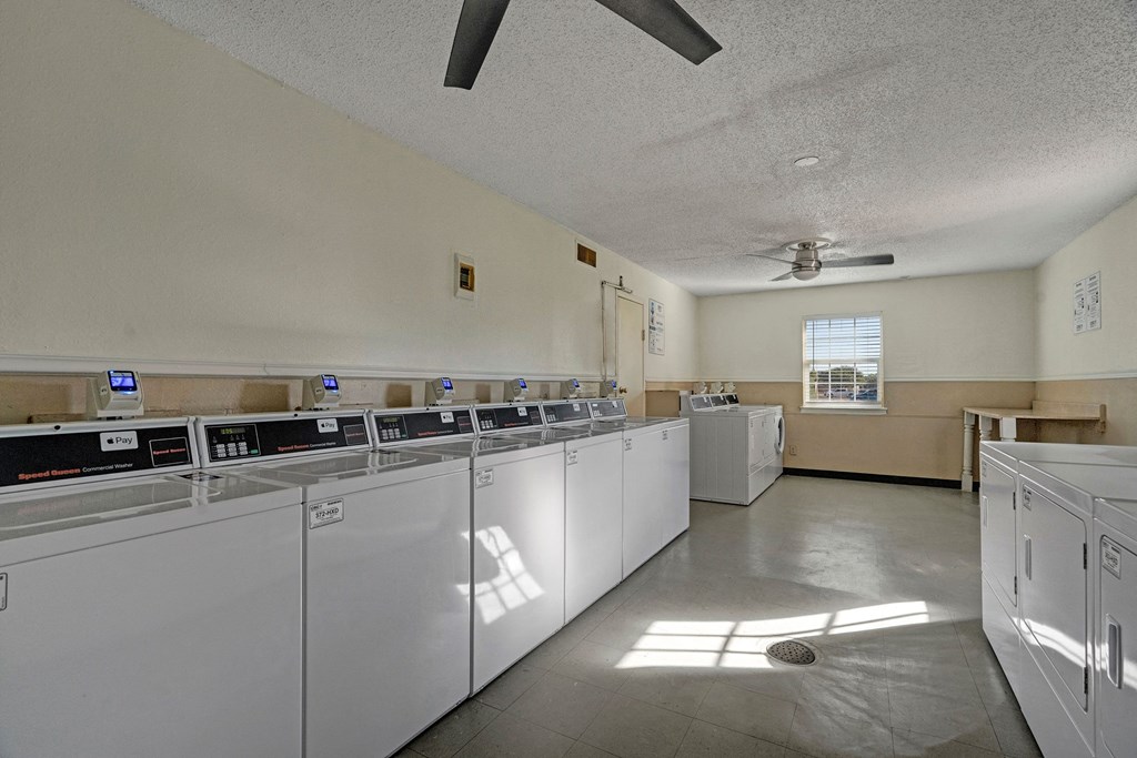 A well-lit laundromat here at Golden Crest Apartment Homes with a row of white washing machines, bright sunlight streaming through a window, casting geometric shadows on the floor.