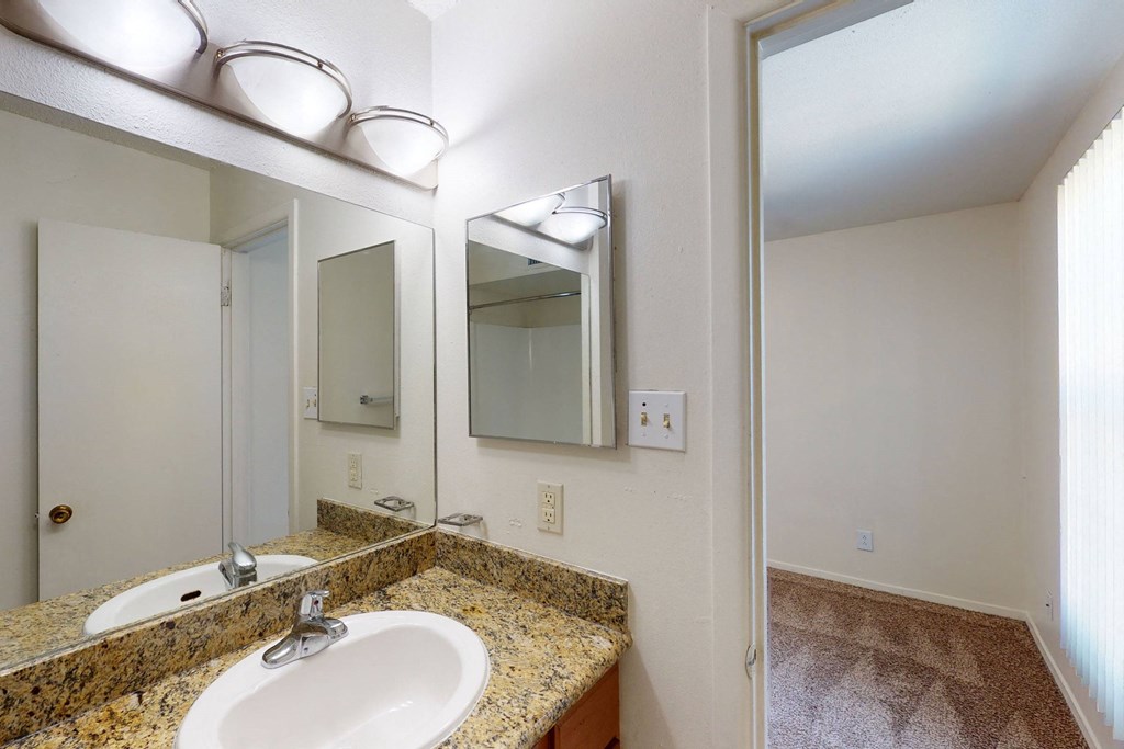 Clean bathroom here at Golden Crest Apartment Homes with granite countertop, oval sink, and rectangular mirror. Bright overhead lighting and an open door leading to a carpeted room.