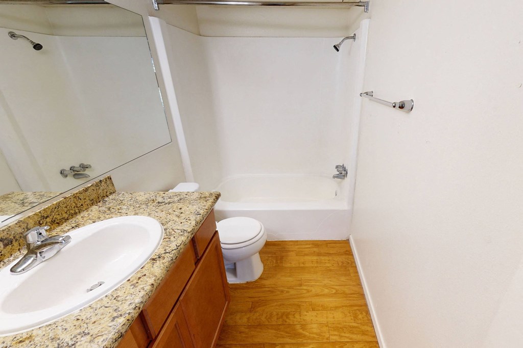 Compact bathroom here at Golden Crest Apartment Homes with a granite countertop and sink, wooden cabinets, and beige walls. Features a white bathtub and toilet on a hardwood floor.