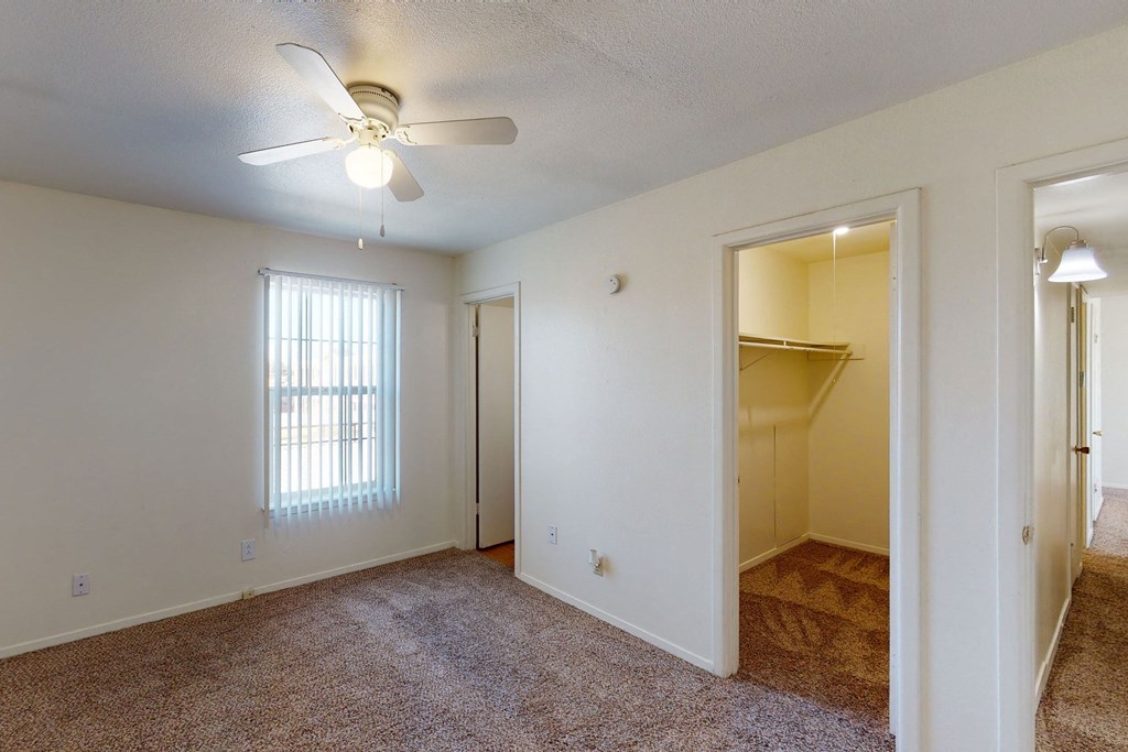 Empty bedroom here at Golden Crest Apartment Homes with beige walls and carpet, featuring a ceiling fan with a light. A window with vertical blinds is on the left, and an open walk-in closet is visible on the right, exuding a calm and simple atmosphere.