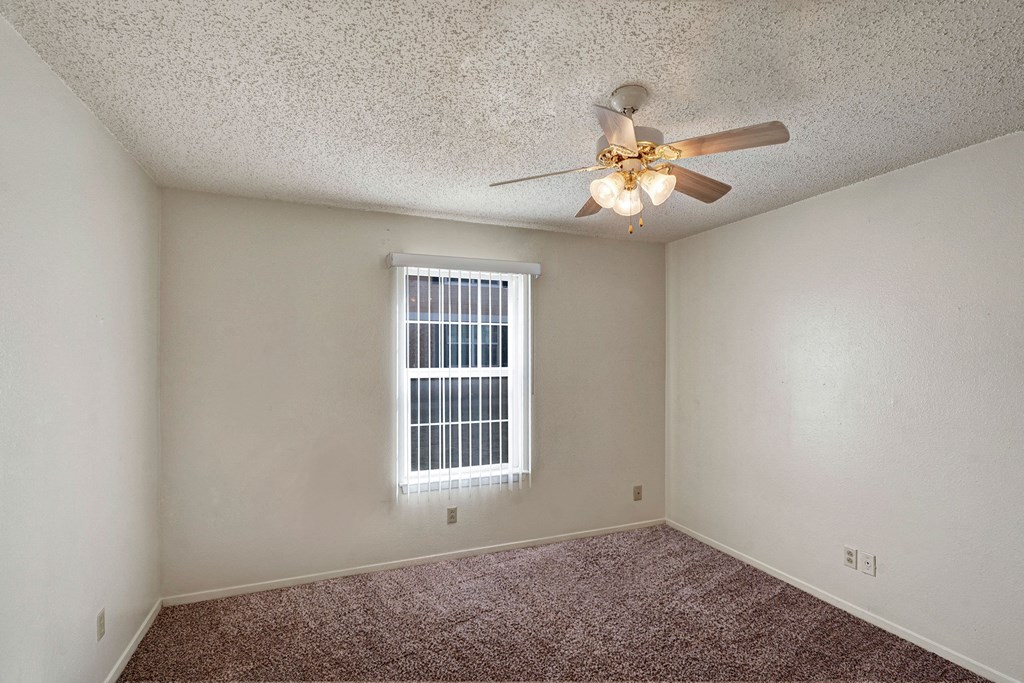Empty bedroom here at Golden Crest Apartment Homes with white walls, brown carpet, a window with vertical blinds, and a ceiling fan with lights, creating a neutral and spacious tone.
