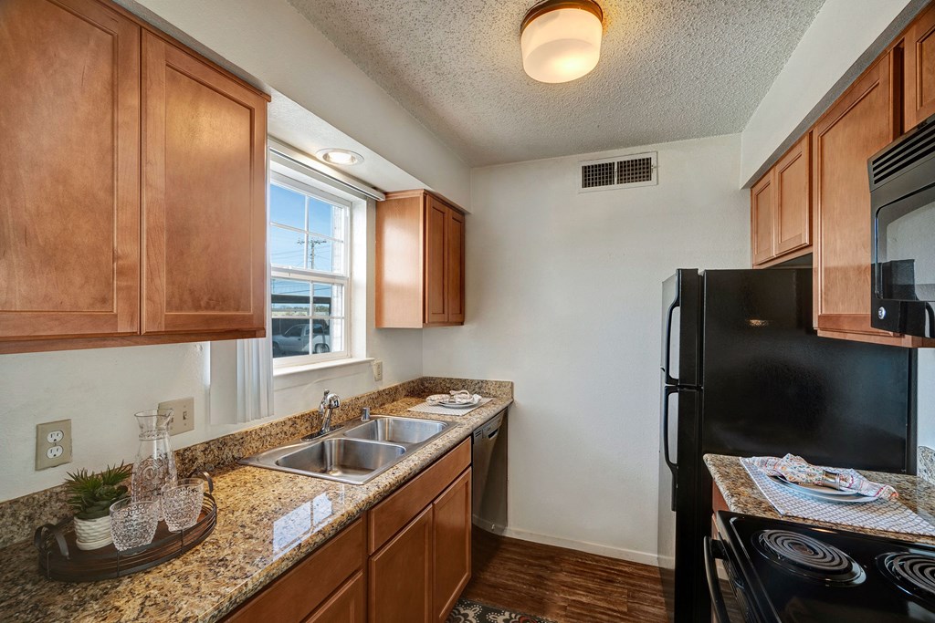 Bright kitchen here at Golden Crest Apartment Homes with wooden cabinets, granite countertops, and stainless steel sink. A black fridge and stove are present, creating a modern feel.