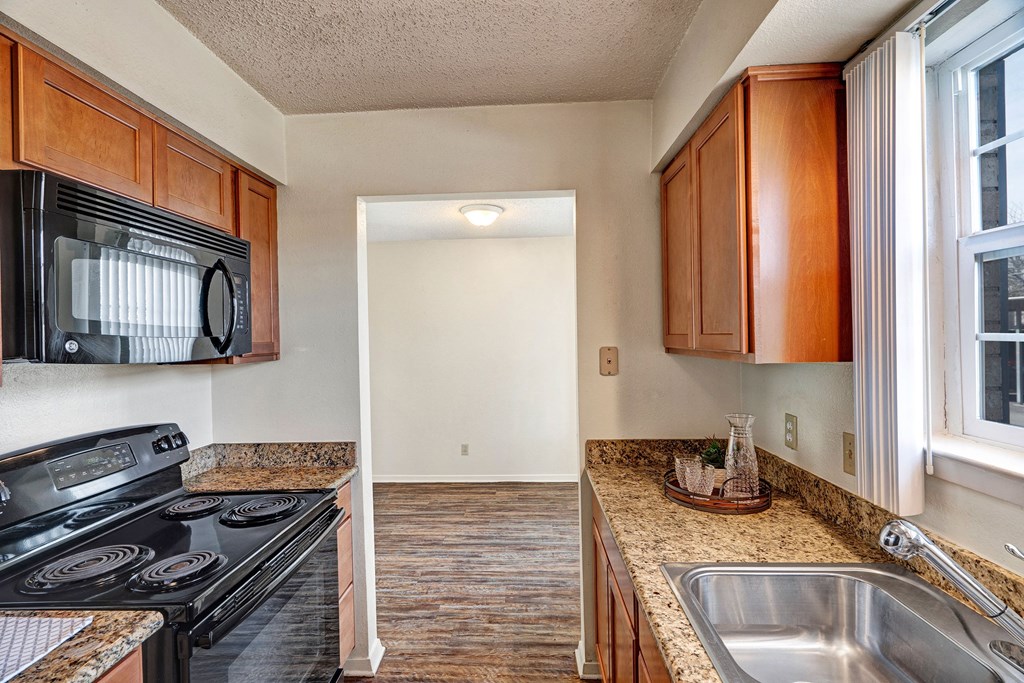Compact kitchen here at Golden Crest Apartment Homes with wooden cabinets, granite countertops, and stainless steel sink. Black stove and microwave on the left, window with blinds on the right.
