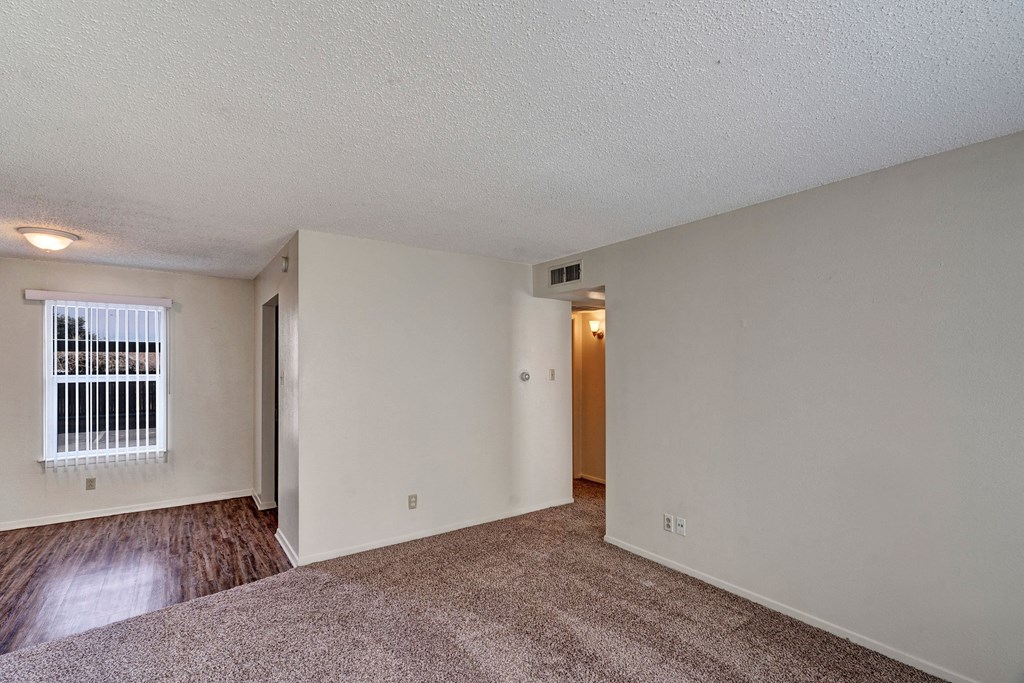 Empty living room here at Golden Crest Apartment Homes with beige walls, carpeted floor, and a transition to wood laminate. A window with vertical blinds lets in natural light. Cozy atmosphere.