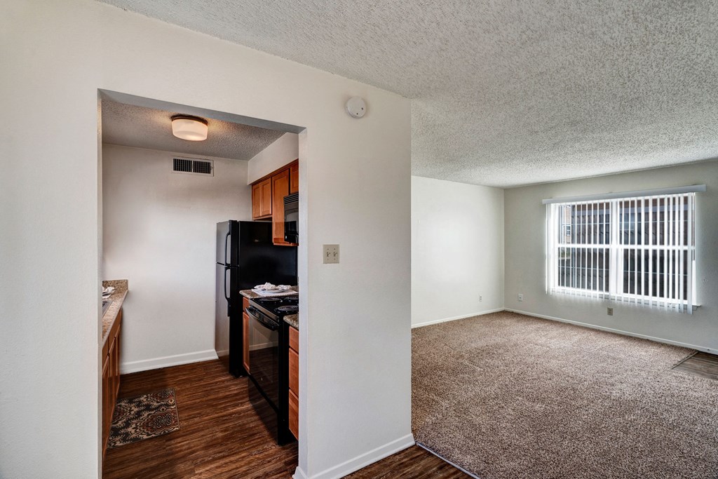 Compact kitchen here at Golden Crest Apartment Homes with black appliances and wooden cabinets opens to a bright, carpeted living room. Large window with blinds, creating a cozy atmosphere.