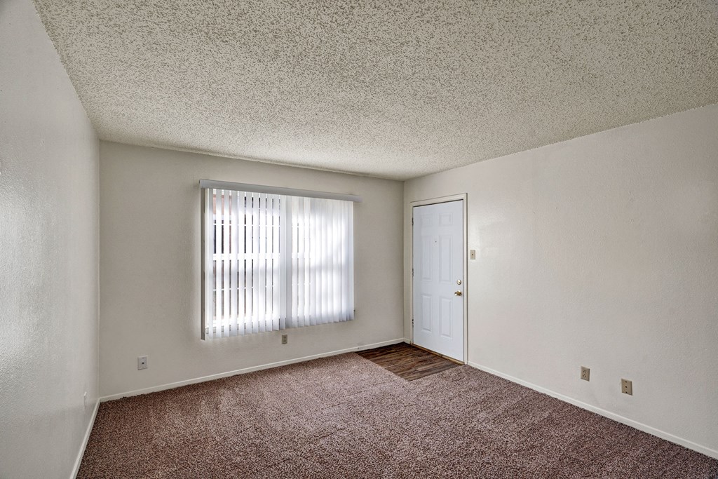 Empty bedroom here at Golden Crest Apartment Homes with beige carpet, white walls, and a textured ceiling. A window with vertical blinds and a white door are on the right, evoking a calm, neutral tone.