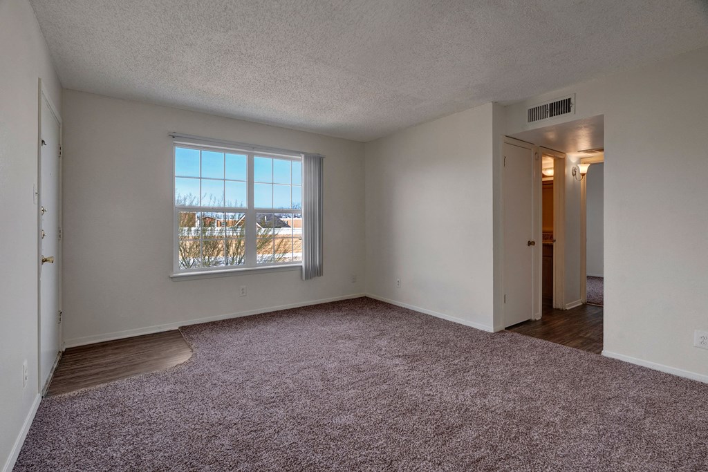 Vacant bedroom here at Golden Crest Apartment Homes with beige walls and carpet, featuring a large window and vertical blinds showing a snowy landscape. Doorway leads to another room. Bright and serene atmosphere.
