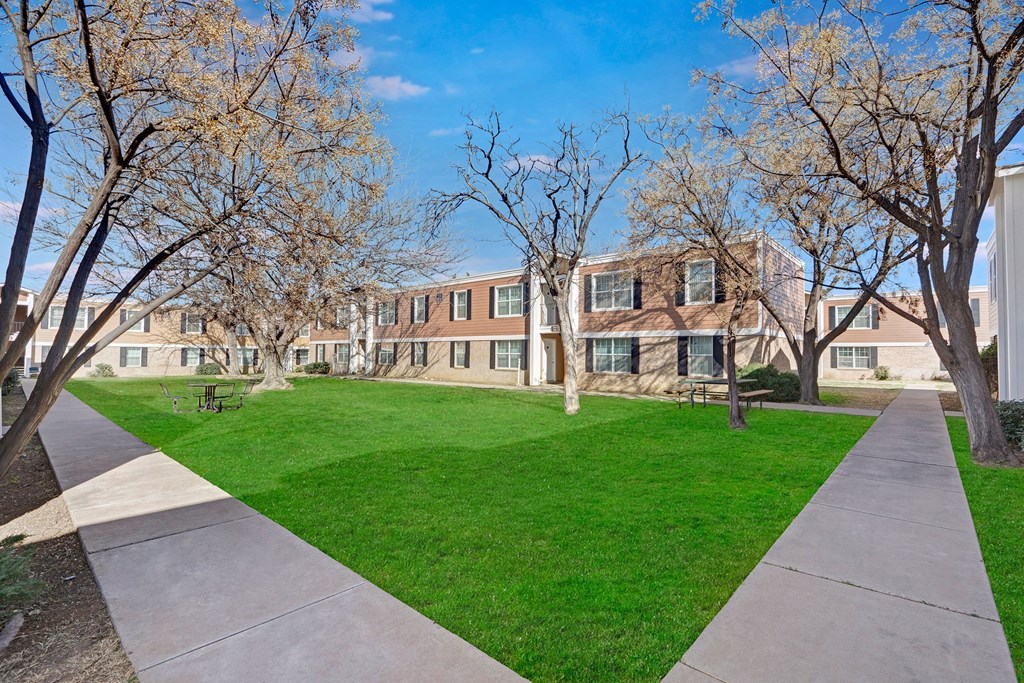 Two-story residential buildings here at Golden Crest Apartment Homes surround a green courtyard with leafless trees. Concrete paths, benches, and a bright, clear sky complete the tranquil scene.