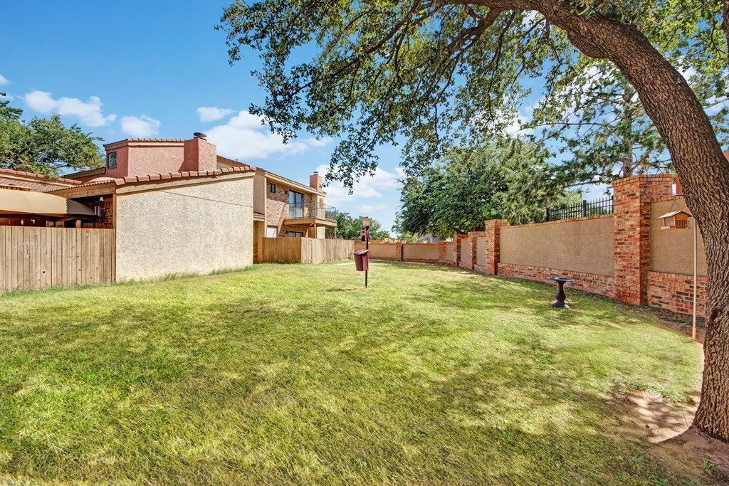 Spacious backyard here at Las Colinas Apartment Homes with green grass, bordered by wooden and brick fences. A tree provides shade, and birdhouses add charm under a clear blue sky.