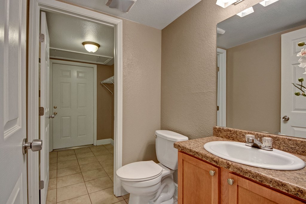 Small bathroom here at Las Colinas Apartment Homes with beige walls and tiled floor, featuring a white toilet and sink with wood cabinets. A mirror reflects a closed door and a lit hallway.