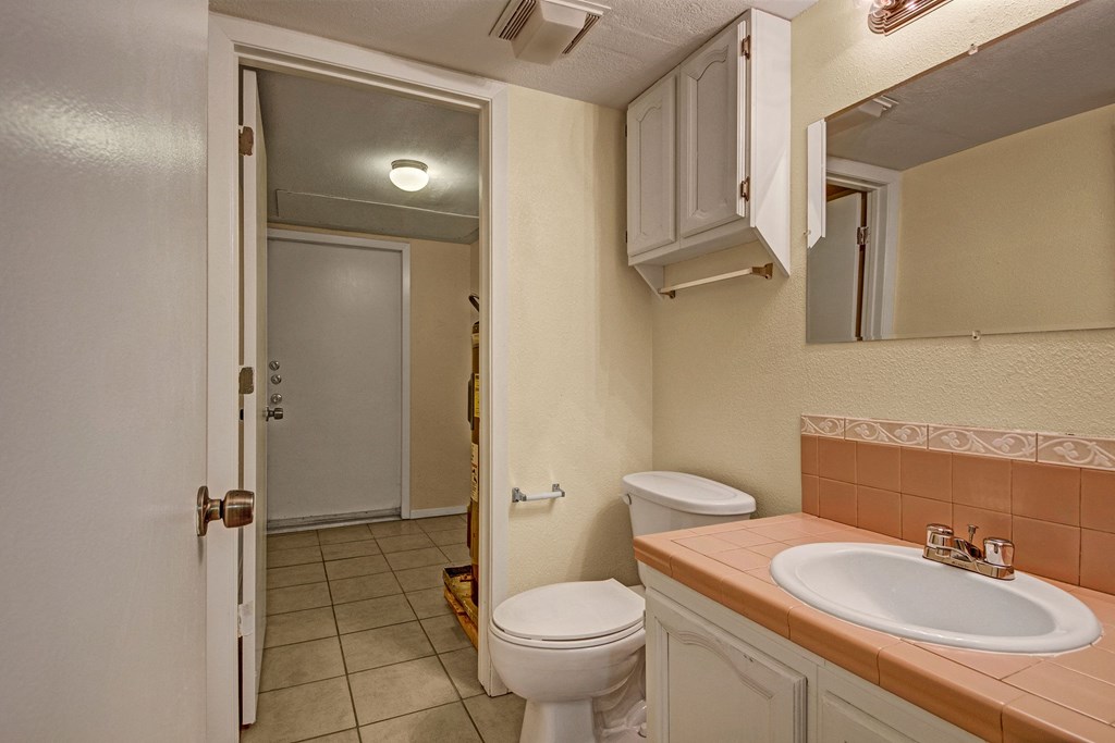 Small bathroom here at Las Colinas Apartment Homes with a tiled floor, peach-colored countertop, and white cabinets. There's a toilet, sink, mirror, and an open door leading to a hallway.
