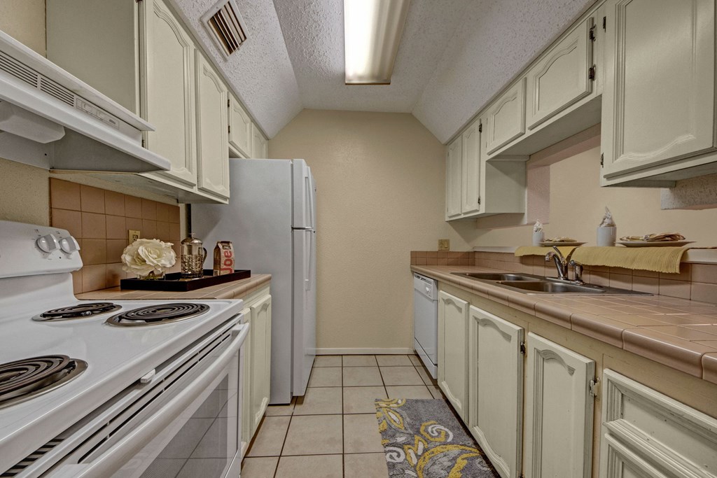 Warm-toned kitchen here at Las Colinas Apartment Homes with white appliances, beige cabinets, and tiled countertops. Decor includes flowers and items on the counter, evoking a cozy feel.