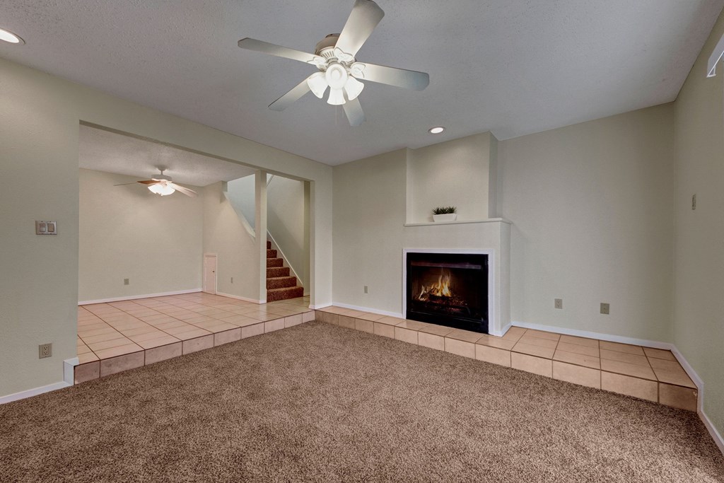 A cozy living room here at Las Colinas Apartment Homes features beige carpet, a lit fireplace, and a ceiling fan. An adjacent tiled area leads to carpeted stairs, creating a warm, inviting atmosphere.