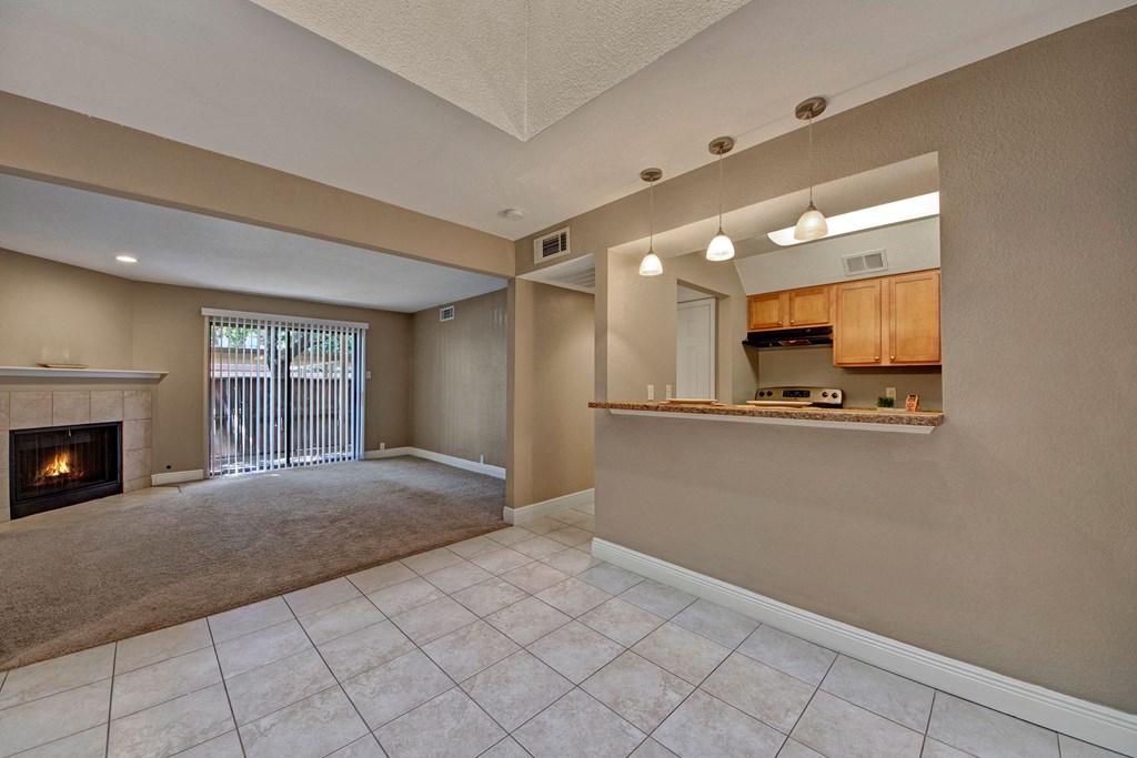 Spacious living area here at Las Colinas Apartment Homes with tiled floor opens to a cozy carpeted space with a fireplace. A kitchen with wooden cabinets and pendant lights is visible.