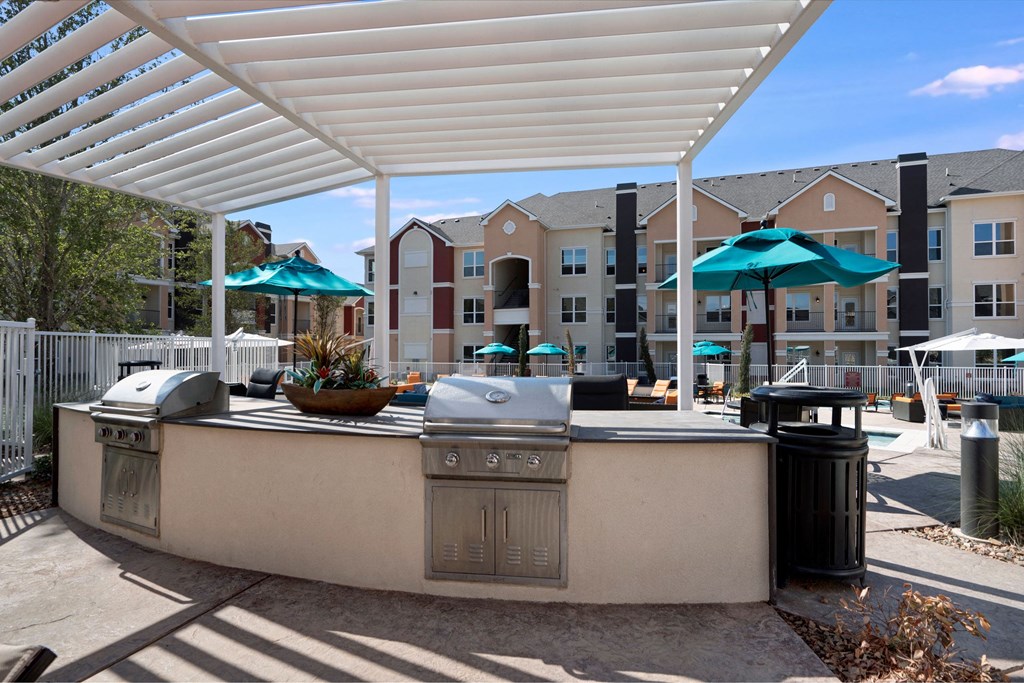 Outdoor patio here at Latitude 31 Apartment Homes with two grills under a white pergola. Background shows apartments, blue sky, and teal umbrellas over tables. Relaxed and inviting ambiance.