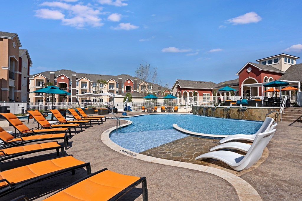 Outdoor pool area here at Latitude 31 Apartment Homes with orange lounge chairs and white reclining chairs beside a clear blue swimming pool. Surrounding red-roofed buildings and umbrellas add a resort-like feel. Sunny weather enhances the vibrant and relaxing atmosphere.