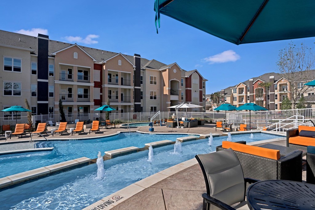 Outdoor pool area here at Latitude 31 Apartment Homes of an apartment complex with fountains, orange lounge chairs, and teal umbrellas. Modern buildings in the background under a clear blue sky.