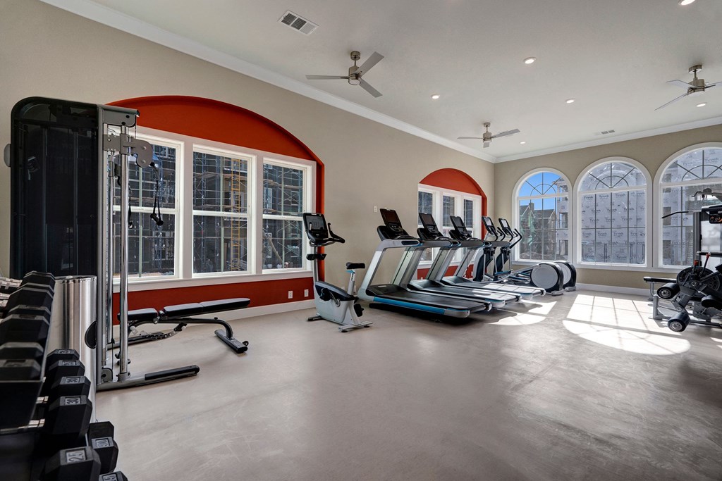 A bright, spacious gym here at Latitude 31 Apartment Homes features treadmills, ellipticals, and strength machines with large arched windows and red accents, conveying a modern, inviting atmosphere.