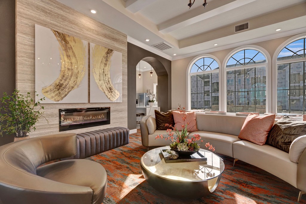 Elegant lounge here at Latitude 31 Apartment Homes with a neutral curved sofa, modern armchairs, and a round coffee table. A chandelier hangs from the coffered ceiling, with artwork and a fireplace adding warmth. Arched windows and doorway accentuate the space.