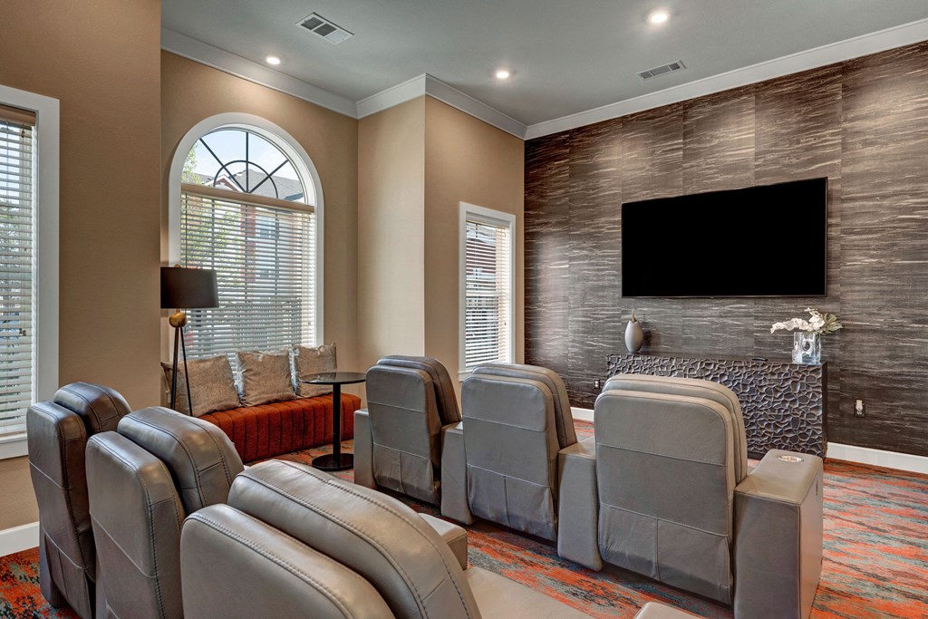 A cozy home theater here at Latitude 31 Apartment Homes with four leather recliners facing a large, wall-mounted flat-screen TV. Soft lighting, modern decor, and warm tones create a relaxing ambiance.