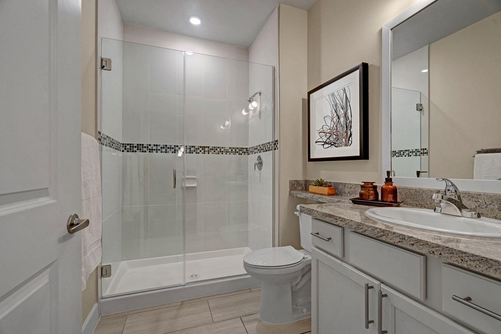 Modern bathroom here at Latitude 31 Apartment Homes with a glass-enclosed shower, mosaic tile accent, and beige walls. A white vanity with a granite countertop, sink, and framed artwork adds elegance.