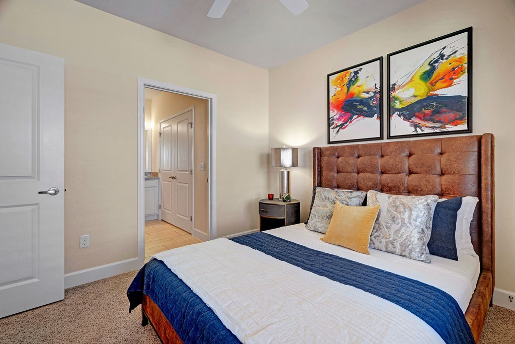 A cozy bedroom here at Latitude 31 Apartment Homes with a brown tufted headboard and colorful pillows on a neatly made bed. Vibrant abstract art hangs on the wall above, adding a lively touch. A bedside table with a modern lamp is next to the bed. An open door reveals a well-lit hallway and bathroom, creating a warm and welcoming ambiance.