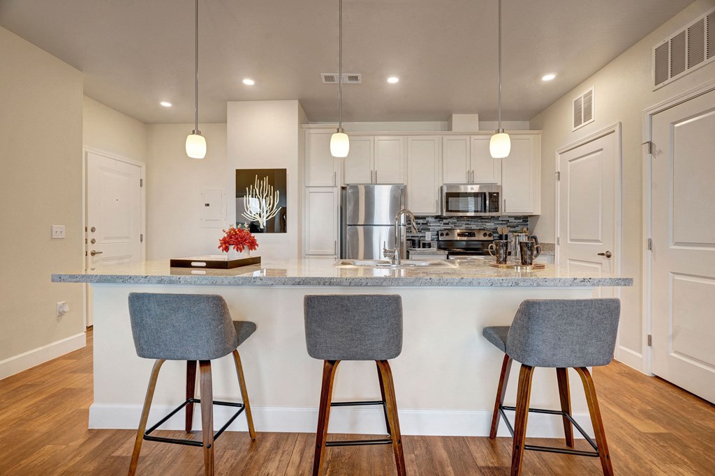 Modern kitchen here at Latitude 31 Apartment Homes with a light gray countertop and three gray barstools. Stainless steel appliances, white cabinets, pendant lights, and hardwood flooring enhance its sleek design.