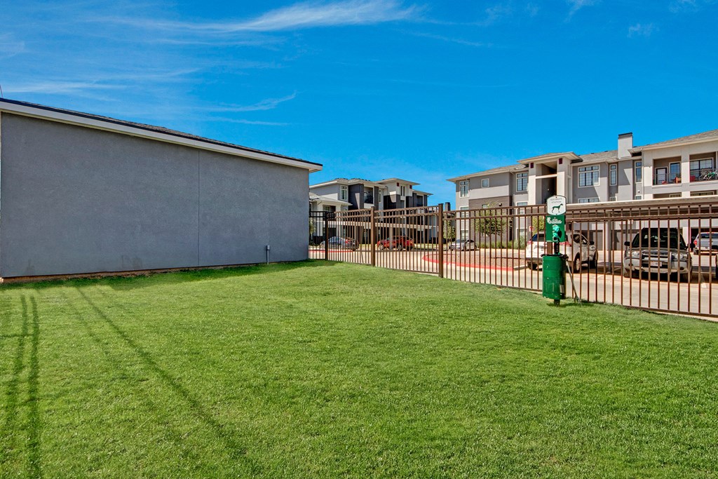 A grassy lawn here at Le Mirage Apartment Homes with a metal fence leads to a modern apartment complex. Clear blue sky enhances the serene, suburban atmosphere.
