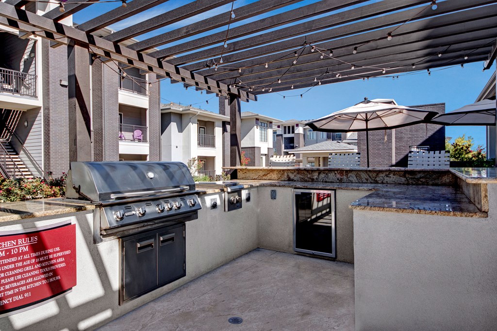 Outdoor patio here at Le Mirage Apartment Homes with a modern grill under a wooden pergola adorned with string lights. Nearby, a granite countertop and a large umbrella offer shade. Residential buildings are visible in the background. The atmosphere is inviting and ready for a gathering.