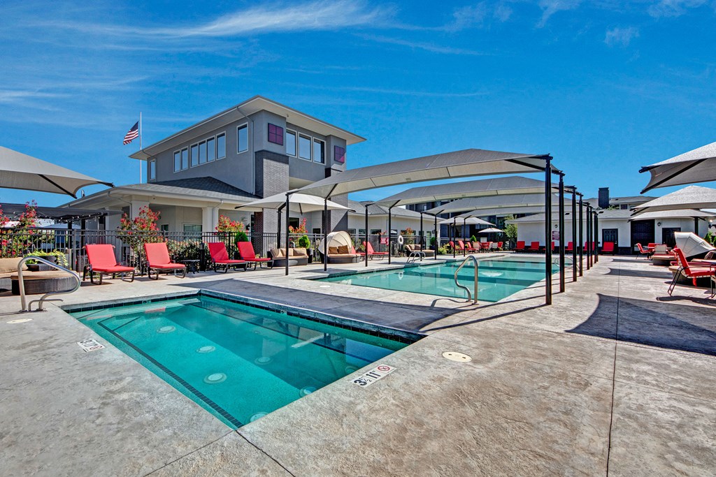 Outdoor pool area here at Le Mirage Apartment Homes with clear blue water, surrounded by red lounge chairs and umbrellas. Modern building in the background with a bright sky above.