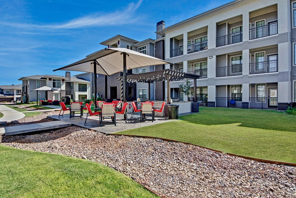 A modern apartment complex here at Le Mirage Apartment Homes with three floors and balconies. In the foreground, a seating area with red chairs and large umbrellas, set on a neatly landscaped lawn. Bright, clear blue sky.