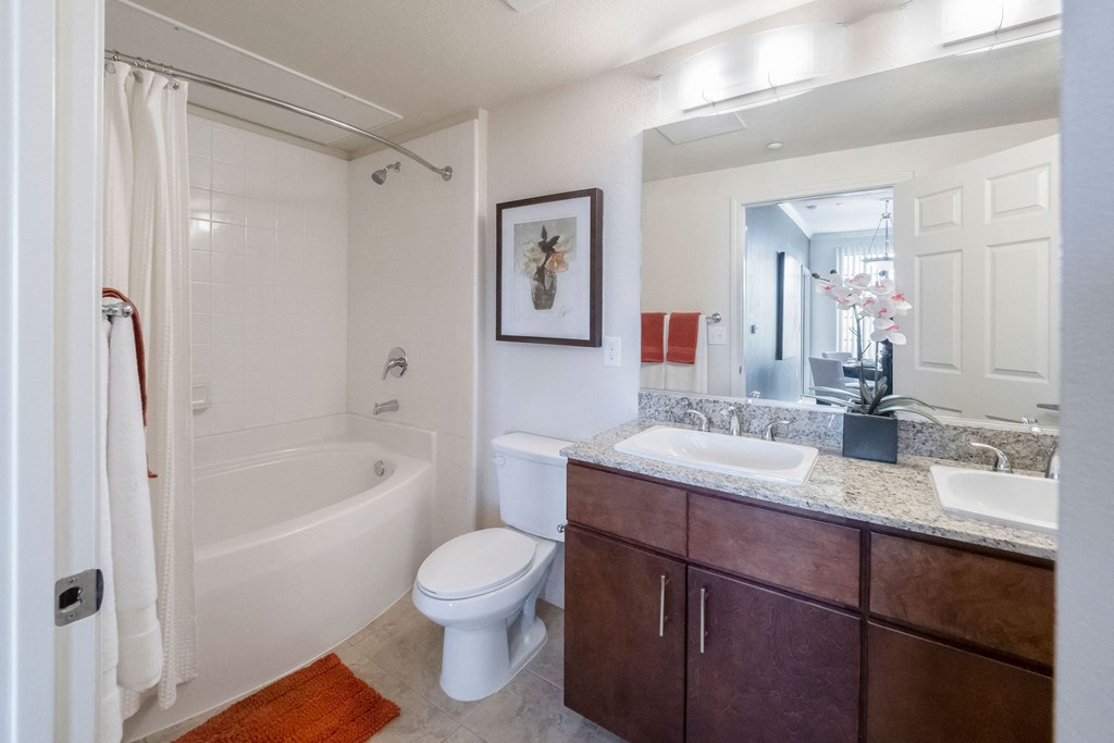 Modern bathroom here at Le Mirage Apartment Homes with a dual-sink vanity, granite countertop, and wooden cabinets. A bathtub with a shower curtain is beside the toilet. Bright and tidy ambiance.