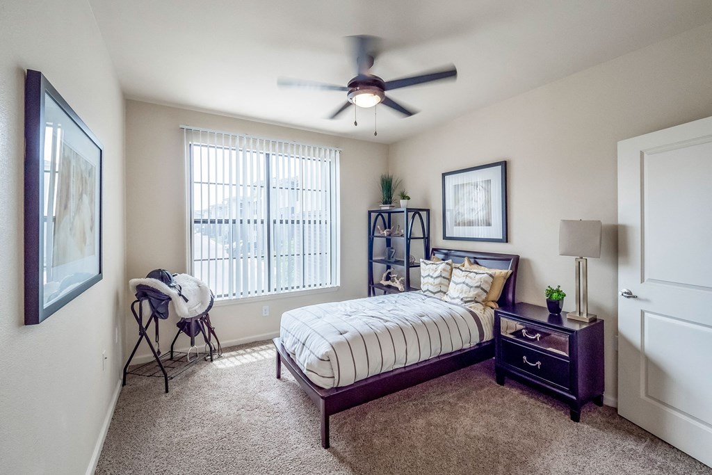Cozy bedroom here at Le Mirage Apartment Homes with modern decor, featuring a bed with striped bedding, a side table with a lamp and plant, a ceiling fan, and a window with blinds.