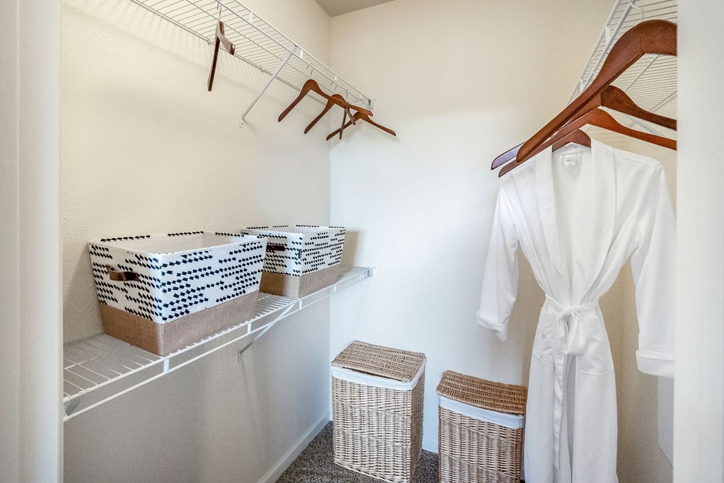 Neatly organized white closet here at Le Mirage Apartment Homes with wire shelves, boxes, wicker baskets, and wooden hangers. A white robe hangs on the right, conveying simplicity.