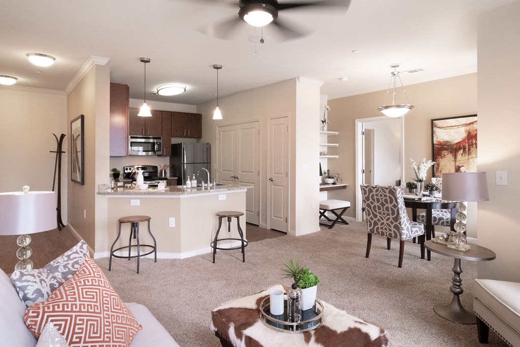 Modern living room here at Le Mirage Apartment Homes with neutral tones, featuring a beige sofa, patterned throw pillows, cowhide ottoman, open kitchen with bar stools, and elegant dining area.