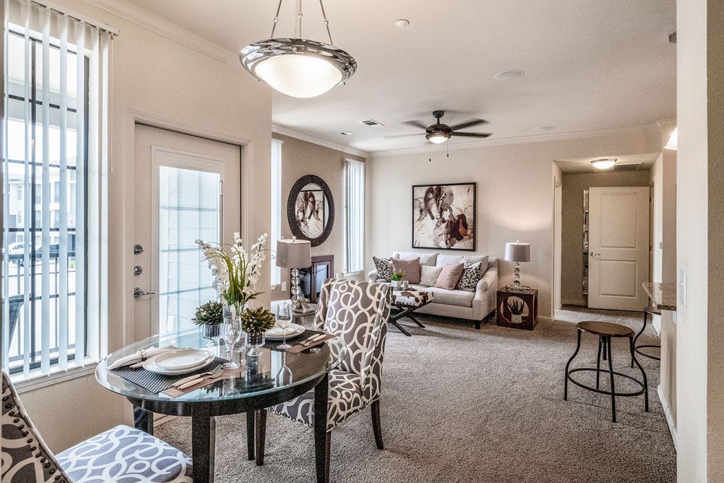 Bright living room here at Le Mirage Apartment Homes with a modern dining area. Features patterned chairs, a glass table, cozy sofa, art, ceiling fan, and large windows on beige walls.