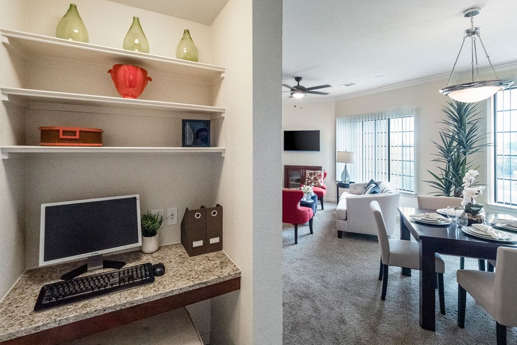 A cozy home office here at Le Mirage Apartment Homes with a computer on a granite desk. Shelves display decorative items. Adjacent is a bright, inviting living-dining area with neutral tones.