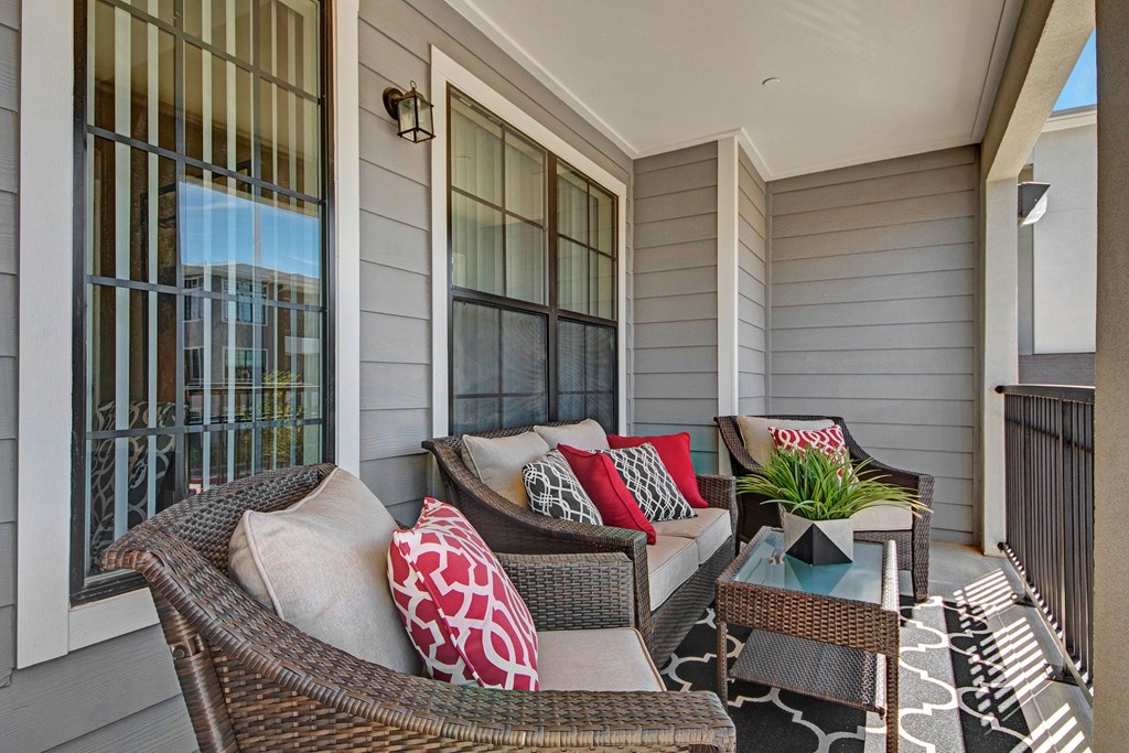 Cozy balcony here at Le Mirage Apartment Homes with wicker furniture, beige cushions, red and patterned pillows, and a small glass table with a plant. Gray siding and a sunny, relaxed vibe.