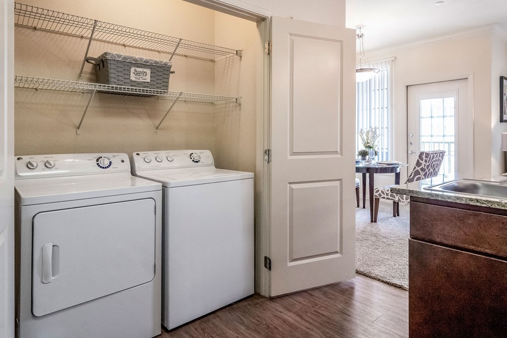 Laundry area here at Le Mirage Apartment Homes with a washer and dryer in a closet with open doors. A shelf above holds a gray basket. Kitchen and dining area visible in the background.