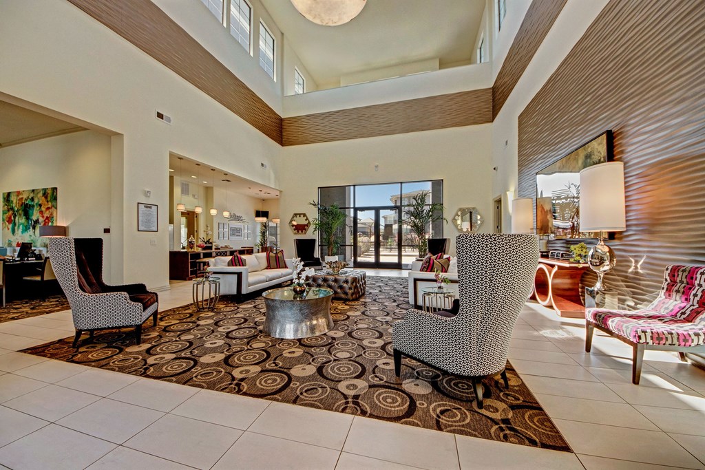 Spacious lobby here at Le Mirage Apartment Homes with high ceilings and modern decor. Includes patterned chairs, round coffee tables, a large rug, and glass doors opening to the outside.