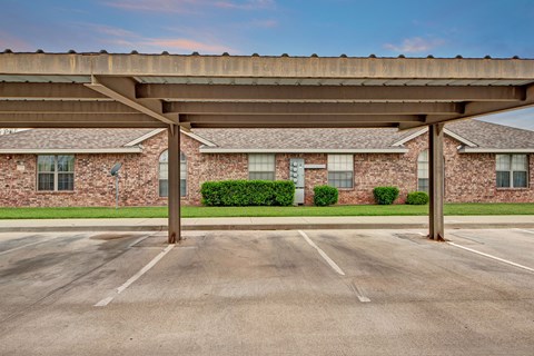 Empty covered parking spaces face a brick building here at Lexington Court Apartment Homes with windows and green shrubs. The scene is calm, with a soft blue sky and hints of evening light.