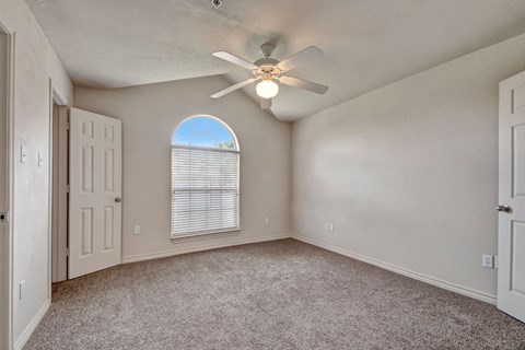 A bright, empty room here at Lexington Court Apartment Homes with beige walls and carpet flooring. It features a ceiling fan with light and an arched window with blinds, creating an airy feel.