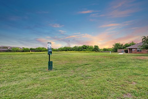 A serene park scene at sunset here at Lexington Court Apartment Homes with a vast grassy field, a dog waste station in the foreground, and a small brick pavilion to the right, under a colorful sky.