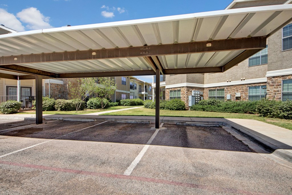 Covered parking area here at Mission Green Apartment Homes with empty spaces in front of an apartment building. Surrounding greenery and blue skies create a calm, suburban setting.