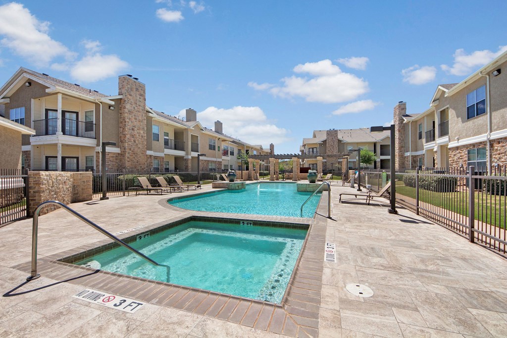 Outdoor pool area here at Mission Green Apartment Homes with beige buildings and balconies. Bright blue sky with scattered clouds, loungers around the poolside.