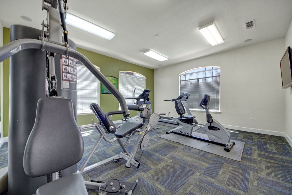 Bright gym room here at Mission Green Apartment Homes with exercise equipment including a weight machine and two stationary bikes. Large windows let in natural light, creating an inviting atmosphere.